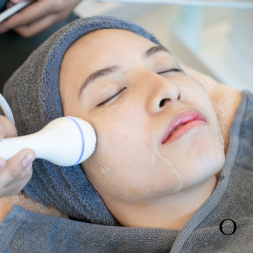 A person is lying down with a cloth on their head, receiving a facial treatment with a device that is applying a product to their face.